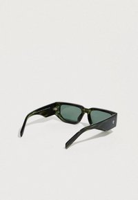 Γυαλιά ηλίου με πράσινη απόχρωση και γεωμετρικό σχήμα, κατασκευασμένα από γυαλιστερό μαύρο υλικό. Διαθέτουν χοντρές βραχίονες και διακριτικές λεπτομέρειες λογοτύπου στο πλευρό.