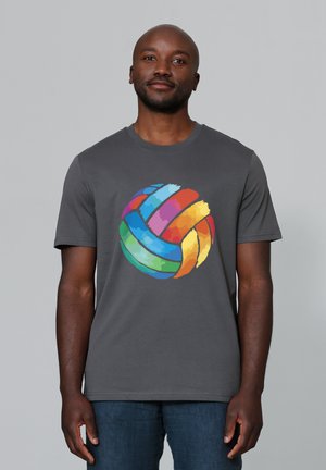 Man draagt een donkergrijs t-shirt met een kleurrijke volleybalafbeelding, staande tegen een effen lichtgrijze achtergrond.