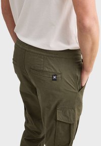 Olijfgroene cargobroek met een comfortabele elastische waistband, zijzakken en een klein zwart logo-opdruk op de achterkant. Katoenen mix stof.