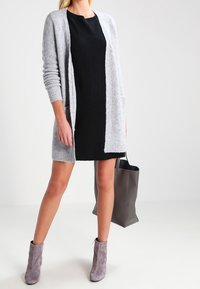 Lång grå cardigan med texturerad stickning, öppen framsidan och två fickor, lagd över en svart klänning. Grå ankelboots och en grå toteväska fulländar looken.