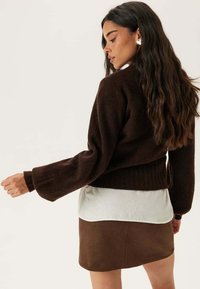 Maglione corto marrone con maniche lunghe, orlo a costine, indossato sopra a una camicia bianca; abbinato a una mini gonna in pelle marrone.