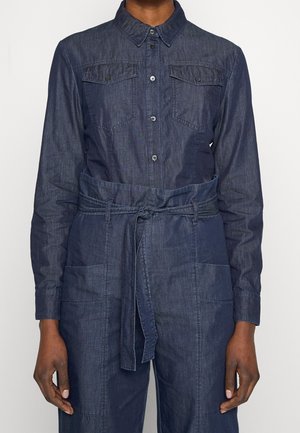Person trägt einen langärmeligen, dunklen Denim-Overall mit Kragen, Knopfleiste, Brusttaschen und einem Stoffgürtel, der in der Taille gebunden ist.