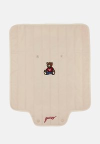 Beige vadderad matta med två tryckknappar, med en broderad björn i röd jacka och blå byxor, samt "guess" logotyp i röd.