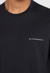 Černé tričko vyrobené z bavlny, s kulatým výstřihem a malým bílým logem "BLACKBARRETT" na levé části hrudi.