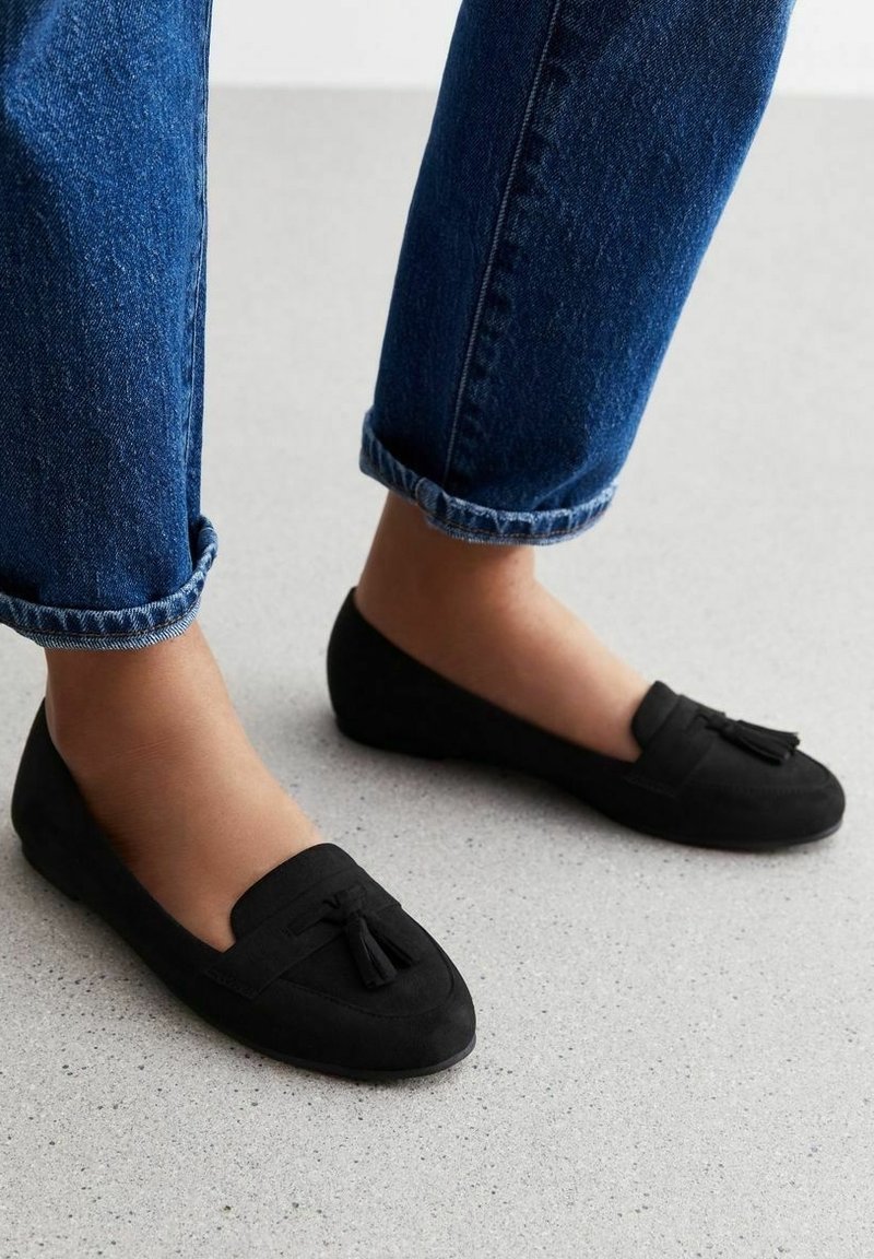 Pieds portant des mocassins en suède noir avec des pampilles, assortis à un jean en denim bleu retroussé sur un sol gris clair.