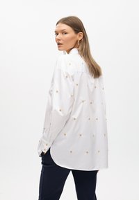 Camicia bianca button-up con dettagli ricamati dorati, maniche lunghe, orlo curvo e vestibilità rilassata, abbinata a pantaloni blu scuro.