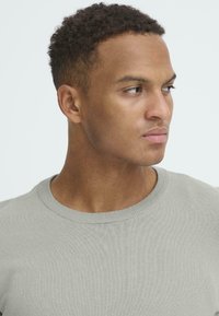 Pulover gri deschis, cu textura ribbed și decolteu rotund, având un croi ajustat și o textură moale. Poziția unghiulară a modelului subliniază forma articolului vestimentar.