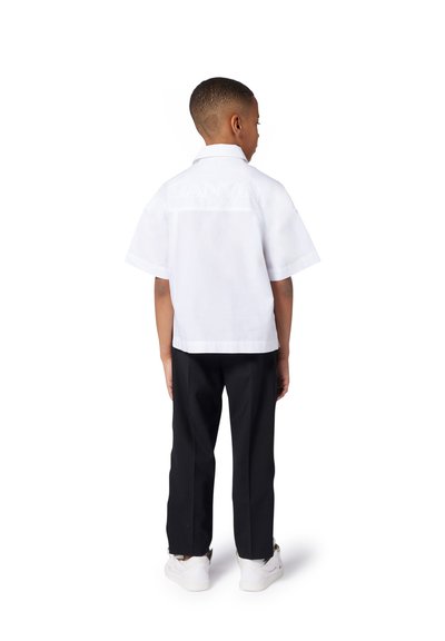 Witte korte mouwen shirt met logodetail op de achterkant, gecombineerd met zwarte broeken en witte sneakers, staande tegen een effen achtergrond.