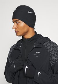 Bonnet de course en tricot noir avec texture c�ôtelée, arborant un logo Nike en argent. Associé à une veste noire et des gants, également dotés d'accents Nike.