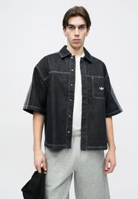 Νεαρός που φοράει μαύρο κοντομάνικο τζιν μπουφάν με λευκά ραφές, γκρι φόρμα, κρατώντας μαύρο καπέλο μπροστά σε μονόχρωμο φόντο.