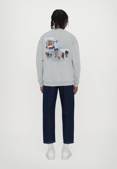 Lichtgrijze sweatshirt met een achterillustratie van een persoon die meerdere honden uitlaat; voorzien van ribgebreide boorden en een zoom, gedragen met donkerblauwe jeans.
