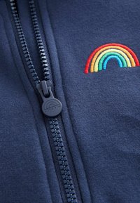 Námornícky zipsový kabát má hladkú textúru, s dúhovou výšivkou nad zipsom a okrúhlym čiernym ťahákom na zips so značkovaním.