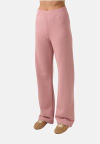 Asmuo, vilkintis aukšto liemens laisvalaikio rožinės spalvos kelnes ir rudus zomšinius sportbačius, ant vienspalvio balto fono.