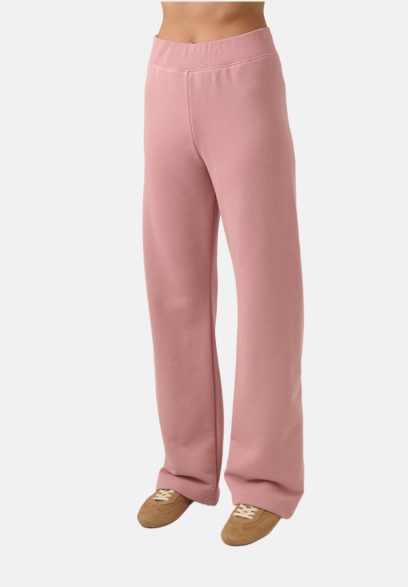 Asmuo, vilkintis aukšto liemens laisvalaikio rožinės spalvos kelnes ir rudus zomšinius sportbačius, ant vienspalvio balto fono.