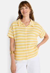 Camisa de manga corta de botones a rayas amarillas y blancas con cuello y tejido texturizado, que presenta un corte relajado y un dobladillo redondeado.