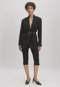 Zwarte op maat gemaakte blazer met diepe V-hals, cinched met een glanzende zwarte riem. Gecombineerd met zwarte capri-legging en zwarte pumps met spitse neus.