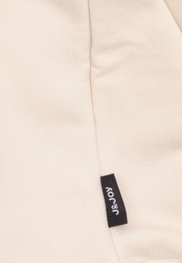 Tissu couleur crème avec une texture côtelée et des détails de couture ; présente une étiquette noire avec du texte blanc, "J&JOY."