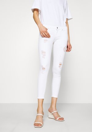 Jeans skinny blancos desgastados con múltiples roturas, con una cintura de tiro medio y un ajuste slim. Combinados con sandalias de cuña blancas.