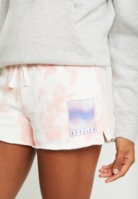 Bílé šortky s jemným růžovým tie-dye vzorem, s modrou grafikou "ATELIER" na levé straně. Měkký materiál a pohodlný volný střih.