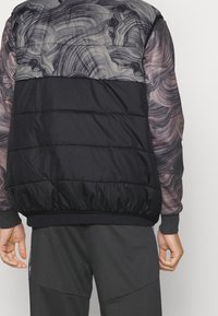 Puffjacka med en svart nedre del, prydd med ett virvlande grått och lila abstrakt mönster på de övre ärmarna och ryggen.