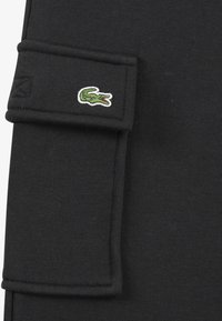 Čierna látka s hladkou textúrou, obsahujúca vrecko a zelené vyšívané logo krokodíla. Minimalistický dizajn s čistými líniami.