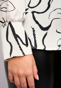 Manche de blouse à motif floral noir et blanc avec un poignet élastique ; le tissu lisse contraste avec des leggings noirs et une main reposant sur le tissu.