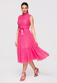 Femme portant une robe en dentelle rose sans manches avec une ceinture ruban et une jupe transparente à pois, posant avec un pied légèrement levé.