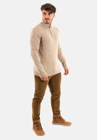 Pull beige à col montant avec fermeture éclair demi-longueur, en tissu tricoté texturé. Associé à un pantalon en velours côtelé marron et des bottes à lacets beiges.