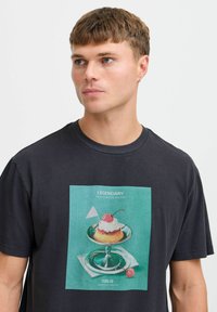Sort bomulds t-shirt med et farverigt print af en dessert på en sølvplade. Designet inkluderer teksten "LEGENDARY" og et kirsebær på toppen.