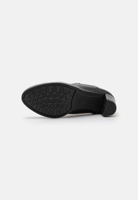 Chaussure en cuir noir inclinée sur le côté, montrant une semelle en caoutchouc à motif et un petit talon empilé sur fond blanc.