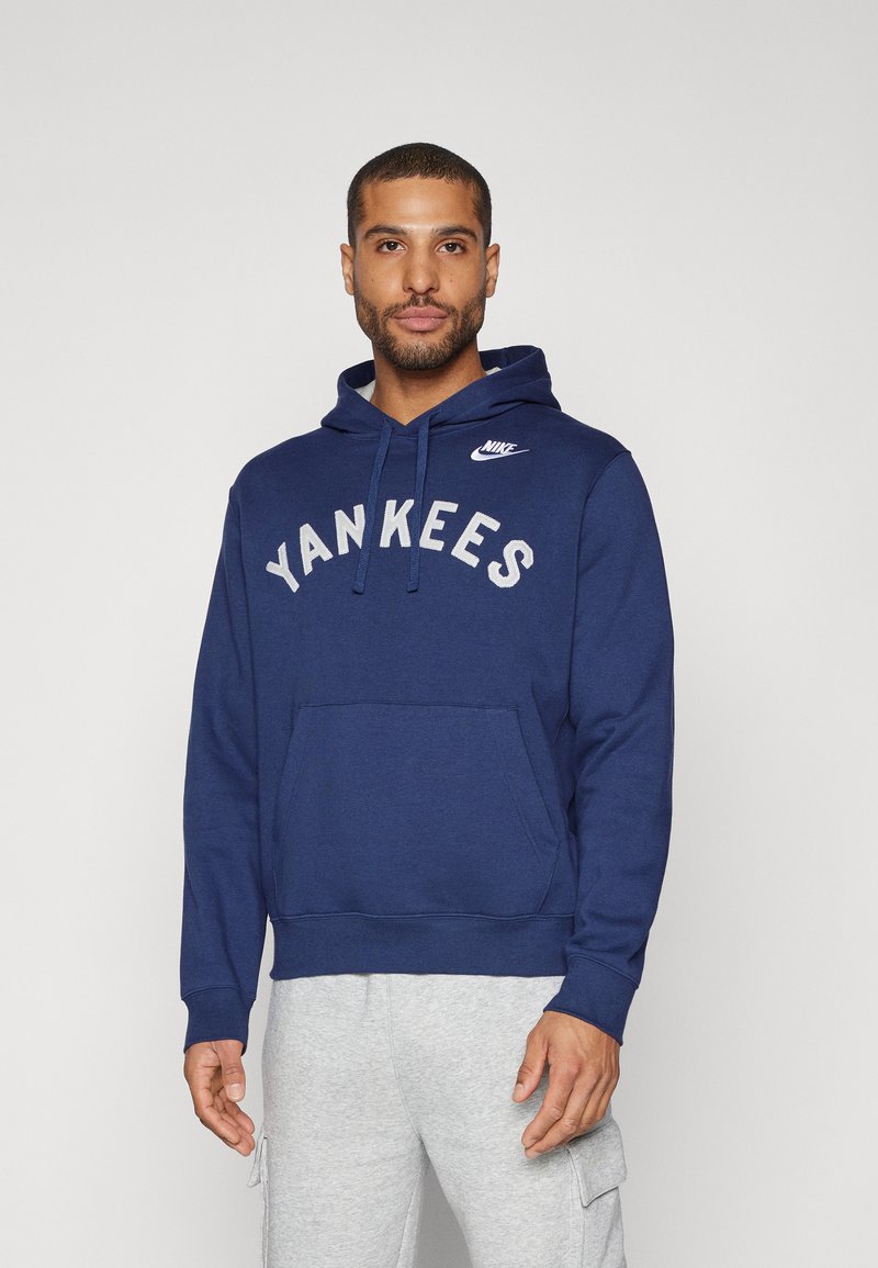 Tumši zils Nike kapučonvīrs ar baltiem burtiem "YANKEES", kangaru kabata, kapuce ar aukliņu un ribotām aprocēm. Pelēki sporta bikses.