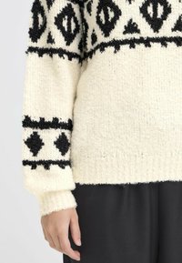 Maglione lavorato a maglia in crema con motivi geometrici neri texturizzati. Presenta polsini e orlo a coste. Abbinato a pantaloni neri larghi.