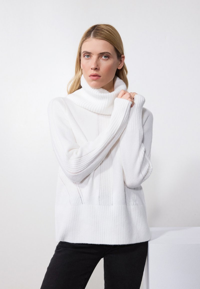 Maglione bianco lavorato a maglia con colletto alto a coste e dettagli in coste verticali. Presenta maniche ampie e un tessuto testurizzato. Abbinato a pantaloni neri.