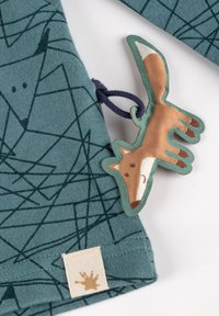 Baumwollstoff in Teal mit Fuchs-Mustern, ausgestattet mit einem plüschigen Fuchs-Tag in Beige und Braun mit einem grünen Rand, befestigt mit einer navyfarbigen Schlaufe.