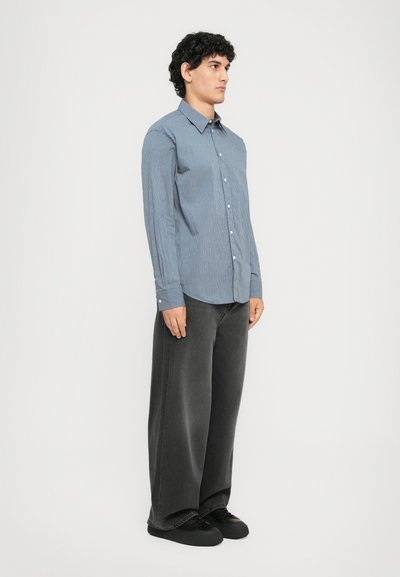 Homme debout tourné vers la droite, portant une chemise à rayures bleues, un jean noir délavé à jambes larges et des baskets noires sur un fond blanc.
