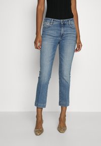 Žena v zkrácených světle modrých džínách, černém tílku bez rukávů a špičatých lodičkách se vzorem hadí kůže stojí na pozadí čistě bílé.