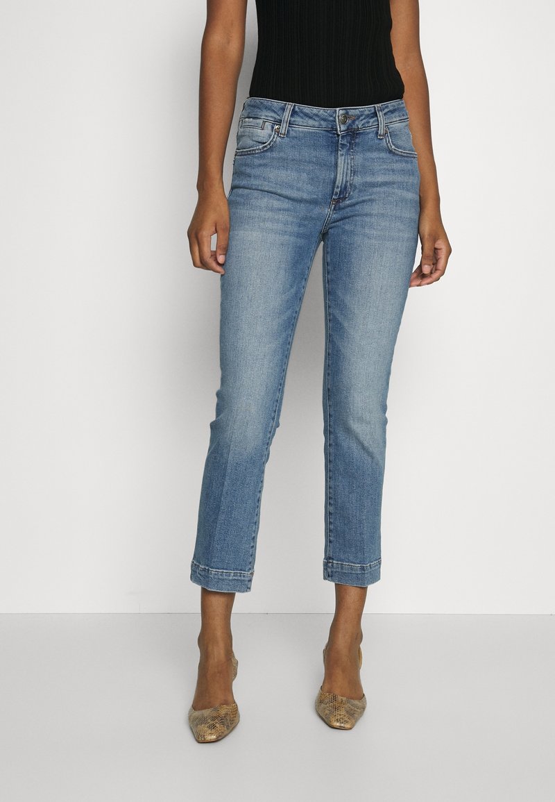 Žena v zkrácených světle modrých džínách, černém tílku bez rukávů a špičatých lodičkách se vzorem hadí kůže stojí na pozadí čistě bílé.