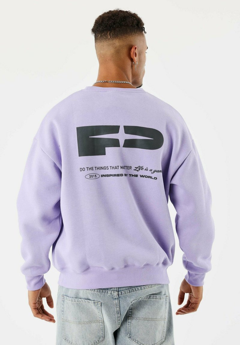 Joven con un sudadera lavanda con el logo negro "FD" y texto en la parte posterior, combinado con jeans de un azul claro, de pie contra un fondo blanco.