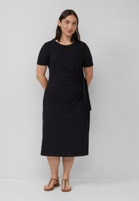Schwarzes Baumwoll-Midi-Kleid mit kurzen Ärmeln, das eine Seitenschnürung in der Taille aufweist. Kombiniert mit hellbraunen Riemchensandalen.