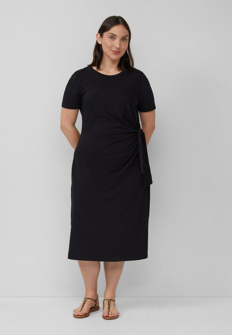 Schwarzes Baumwoll-Midi-Kleid mit kurzen Ärmeln, das eine Seitenschnürung in der Taille aufweist. Kombiniert mit hellbraunen Riemchensandalen.