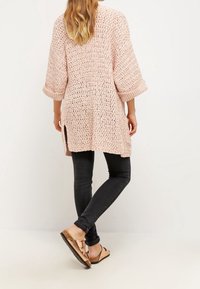 Kobieta stoi, ubrana w luźny różowy sweter z dzianiny, czarne rurki i metaliczne sandały w kolorze różowego złota z dwiema paskami.