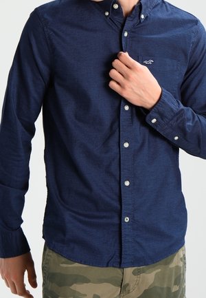 Man wearing a navy blue button-up shirt with a small white logo on the chest, and camouflage-patterned pants.