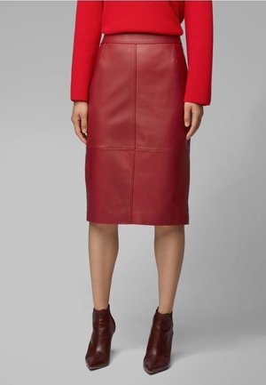 Femme portant une jupe crayon en cuir rouge au genou, un pull rouge à manches longues et des bottines rouge foncé, debout devant un fond gris.