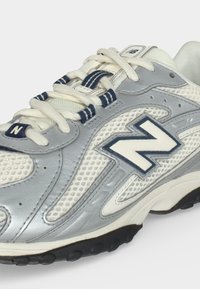 Λευκό και γκρι παπούτσι New Balance με πλέγμα, κορδόνια περασμένα μέσα από θηλιές με ναυτικό ριγέ σχέδιο και μαύρη λαστιχένια σόλα.