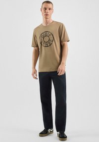 T-shirt en coton couleur beige avec un graphisme circulaire noir portant l'inscription "QUITE SENSE." Associé à un pantalon bleu foncé et des baskets noires avec des accents blancs.