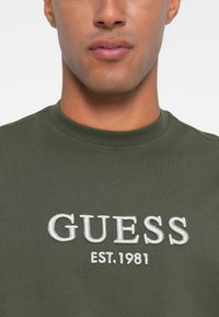 Groene katoenen sweatshirt met ronde hals met daarop geborduurde witte "GUESS" en "EST. 1981" branding aan de voorkant. Gladde textuur, normale pasvorm.