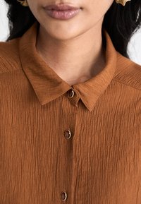 Brown textured shirt with a collar, featuring matching buttons and small gold accents. The fabric has a creased appearance.