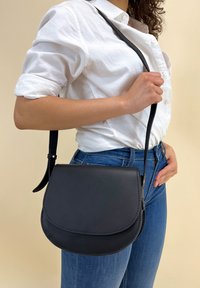 Sort læder crossover-taske med en rund klap, glat tekstur og stikninger langs kanten. Justerbar rem fastgjort til en kompakt silhuet.