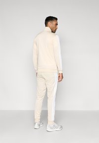 Σετ φόρμας σε κρεμ χρώμα με ψηλό γιακά και λευκές πλαϊνές ρίγες. Κατασκευασμένο από μαλακό υλικό, με εφαρμοστά μανίκια και τελείωμα. Συμπεριλαμβάνονται γκρι αθλητικά παπούτσια.