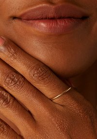 Gold ring on a finger, with textured brown skin in the background. Full lips with a muted lip color are visible, showing subtle curves and details.